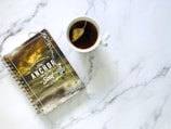 A spiral-bound notebook with a religious theme featuring a stormy sea and a Bible verse on the cover is placed next to a white cup containing tea with a teabag. The notebook has a decorative cross keychain attached.