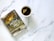 A spiral-bound notebook with a religious theme featuring a stormy sea and a Bible verse on the cover is placed next to a white cup containing tea with a teabag. The notebook has a decorative cross keychain attached.