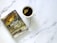 A spiral-bound notebook with a religious theme featuring a stormy sea and a Bible verse on the cover is placed next to a white cup containing tea with a teabag. The notebook has a decorative cross keychain attached.