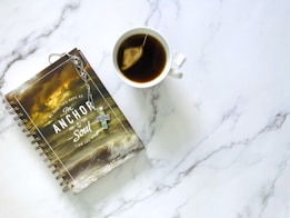 A spiral-bound notebook with a religious theme featuring a stormy sea and a Bible verse on the cover is placed next to a white cup containing tea with a teabag. The notebook has a decorative cross keychain attached.