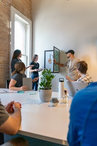 A group of people are engaged in a casual meeting or brainstorming session in a bright room with a large window. One person is standing and pointing at a framed picture on the wall, while others are seated around a table, focusing on the discussion. There is a potted plant on the table, along with various office supplies and personal items.