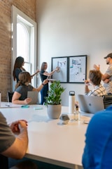 A group of people is engaged in a meeting or brainstorming session in an office environment. Two individuals stand by a wall-mounted board with charts and images, presenting information to the others who are seated around a table. The setting includes a large white table with notepads, a laptop, a water bottle, a small plant, and some stationery. Natural light filters through a nearby window, creating a bright and open atmosphere.
