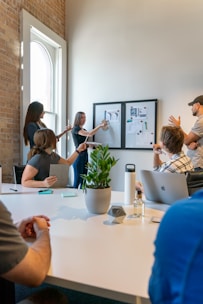 A group of people is engaged in a meeting or brainstorming session in an office environment. Two individuals stand by a wall-mounted board with charts and images, presenting information to the others who are seated around a table. The setting includes a large white table with notepads, a laptop, a water bottle, a small plant, and some stationery. Natural light filters through a nearby window, creating a bright and open atmosphere.