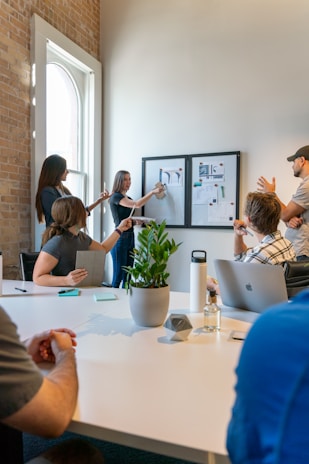 A group of people is engaged in a meeting or brainstorming session in an office environment. Two individuals stand by a wall-mounted board with charts and images, presenting information to the others who are seated around a table. The setting includes a large white table with notepads, a laptop, a water bottle, a small plant, and some stationery. Natural light filters through a nearby window, creating a bright and open atmosphere.
