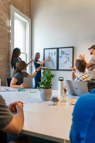 A group of people is engaged in a meeting or brainstorming session in an office environment. Two individuals stand by a wall-mounted board with charts and images, presenting information to the others who are seated around a table. The setting includes a large white table with notepads, a laptop, a water bottle, a small plant, and some stationery. Natural light filters through a nearby window, creating a bright and open atmosphere.