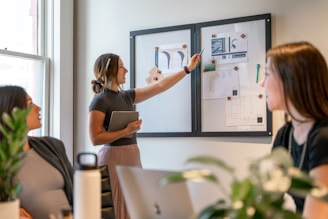 A group of people are engaged in a collaborative meeting in an office setting. A woman stands pointing at a whiteboard with drawings and notes while holding a tablet. Two other women are seated, facing the standing woman, and appear to be attentively listening. The room is well-lit through a large window, and there are some plants and office supplies visible on the table.