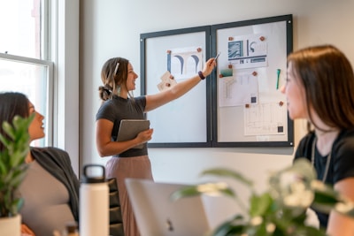 A group of people are engaged in a collaborative meeting in an office setting. A woman stands pointing at a whiteboard with drawings and notes while holding a tablet. Two other women are seated, facing the standing woman, and appear to be attentively listening. The room is well-lit through a large window, and there are some plants and office supplies visible on the table.