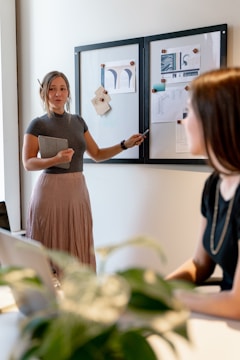 A woman is standing and gesturing towards a whiteboard with various papers attached to it while another woman sits and listens. The setting appears to be a modern office or meeting room. A plant is visible in the foreground, adding a touch of greenery to the space.