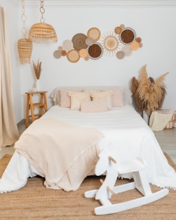 A cozy bedroom with a white bed adorned with beige and cream pillows and a soft blanket. Above the bed is a decorative wall arrangement featuring round, woven textures in various shades of brown and beige. Beside the bed is a wooden stool with a white vase and dry grasses. Pendant lights made from natural materials hang from the ceiling. A small white wooden rocking horse sits on a textured rug.