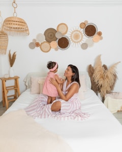 A woman is sitting on a neatly made bed, holding a small child closely. The room has a natural, bohemian decor with woven straw hats and pampas grass. The bed is draped with a light-colored striped sheet, and there are decorative pillows. The walls are adorned with circular woven baskets in various shades.