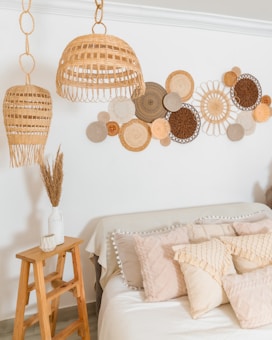 A cozy room with woven rattan pendant lights hanging from the ceiling. A wall decoration made of circular, woven natural fiber designs is displayed. A bed is adorned with textured, cream-colored pillows and a knit throw. A small wooden ladder serves as a table, holding a ceramic vase with dried grasses and a candle.