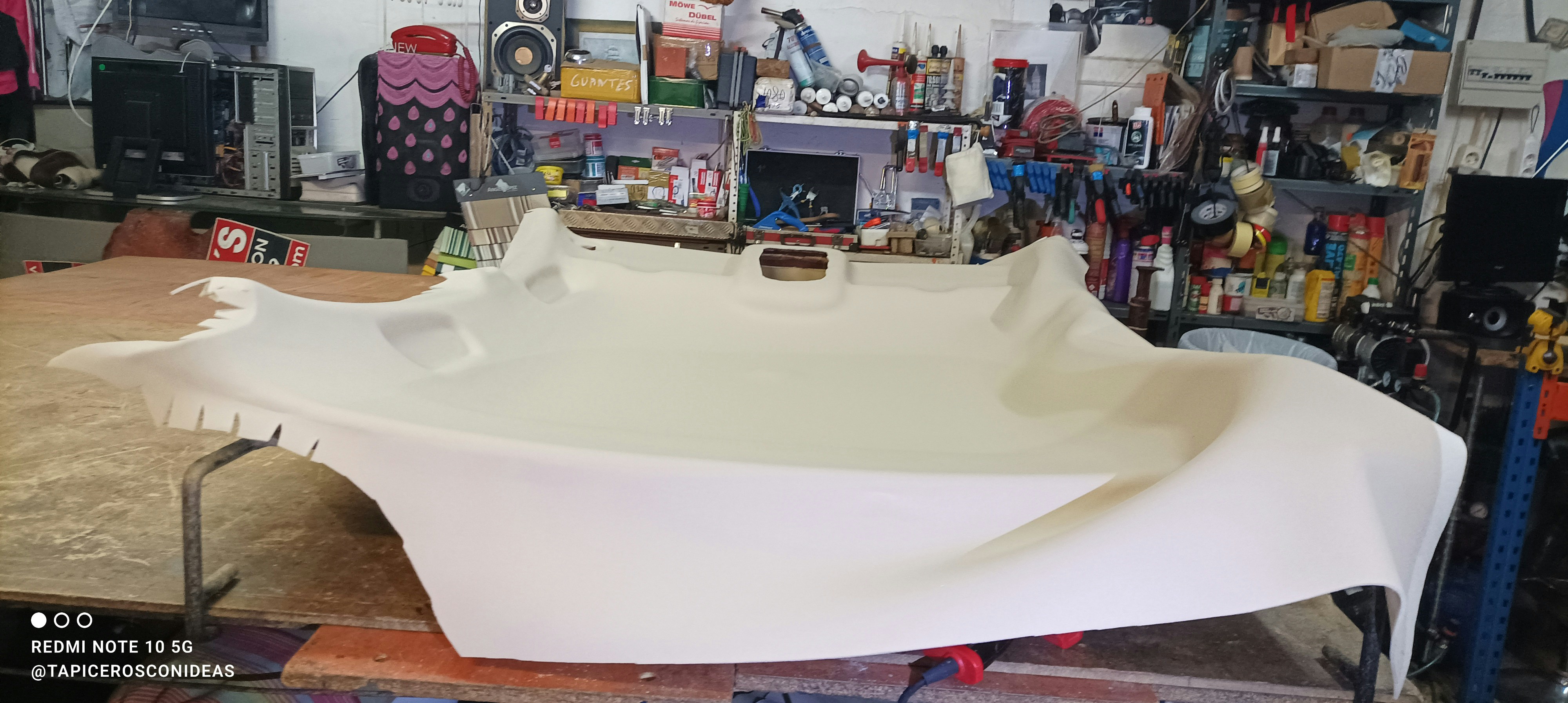 A large piece of beige fabric is spread over a table in a workshop filled with various tools and equipment. The table appears to be used for upholstery work, and the background shows shelves stocked with containers and a variety of mechanical or electronic items.