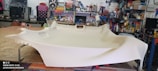 A large piece of beige fabric is spread over a table in a workshop filled with various tools and equipment. The table appears to be used for upholstery work, and the background shows shelves stocked with containers and a variety of mechanical or electronic items.