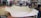 A large piece of beige fabric is spread over a table in a workshop filled with various tools and equipment. The table appears to be used for upholstery work, and the background shows shelves stocked with containers and a variety of mechanical or electronic items.