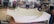 A large piece of beige fabric is spread over a table in a workshop filled with various tools and equipment. The table appears to be used for upholstery work, and the background shows shelves stocked with containers and a variety of mechanical or electronic items.