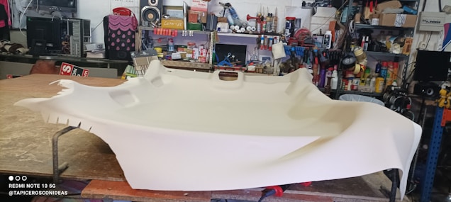 A large piece of beige fabric is spread over a table in a workshop filled with various tools and equipment. The table appears to be used for upholstery work, and the background shows shelves stocked with containers and a variety of mechanical or electronic items.