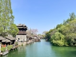 A scenic riverside view featuring a traditional Chinese building with intricate roofs on the left side, surrounded by lush green trees. A calm river runs through the scene, bordered by a stone pathway. Small boats are floating on the water, and the sky is clear and blue.