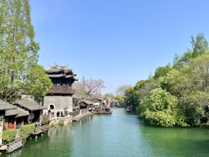 A scenic riverside view featuring a traditional Chinese building with intricate roofs on the left side, surrounded by lush green trees. A calm river runs through the scene, bordered by a stone pathway. Small boats are floating on the water, and the sky is clear and blue.