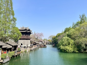 A scenic riverside view featuring a traditional Chinese building with intricate roofs on the left side, surrounded by lush green trees. A calm river runs through the scene, bordered by a stone pathway. Small boats are floating on the water, and the sky is clear and blue.