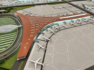 A scale model of an airport terminal featuring a large, brown, V-shaped building with numerous parked airplanes lined up along the terminal gates. The surrounding area includes a series of roads, greenery, and a circular section with multiple lanes and vehicles.