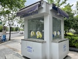 Small white kiosk with windows and circular openings for handling objects without direct contact. The kiosk is situated outdoors on a paved sidewalk with greenery, trees, and other urban elements surrounding it. There are some signs in an Asian language, a couple of blue bicycles, and a vehicle parked on a road in the background.