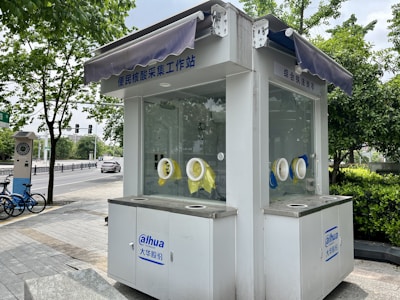 Small white kiosk with windows and circular openings for handling objects without direct contact. The kiosk is situated outdoors on a paved sidewalk with greenery, trees, and other urban elements surrounding it. There are some signs in an Asian language, a couple of blue bicycles, and a vehicle parked on a road in the background.