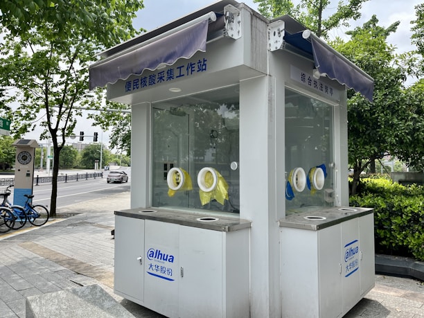 Small white kiosk with windows and circular openings for handling objects without direct contact. The kiosk is situated outdoors on a paved sidewalk with greenery, trees, and other urban elements surrounding it. There are some signs in an Asian language, a couple of blue bicycles, and a vehicle parked on a road in the background.