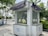 Small white kiosk with windows and circular openings for handling objects without direct contact. The kiosk is situated outdoors on a paved sidewalk with greenery, trees, and other urban elements surrounding it. There are some signs in an Asian language, a couple of blue bicycles, and a vehicle parked on a road in the background.