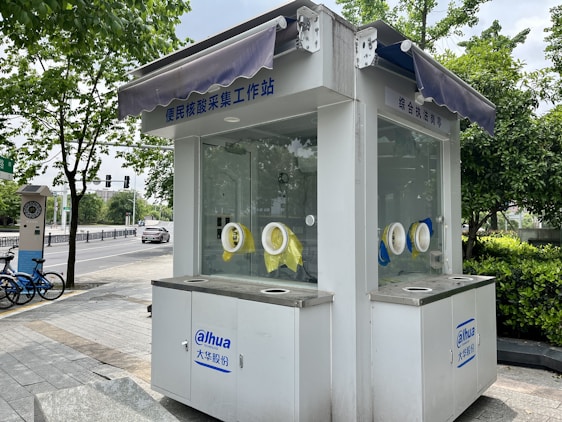 Small white kiosk with windows and circular openings for handling objects without direct contact. The kiosk is situated outdoors on a paved sidewalk with greenery, trees, and other urban elements surrounding it. There are some signs in an Asian language, a couple of blue bicycles, and a vehicle parked on a road in the background.
