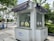 Small white kiosk with windows and circular openings for handling objects without direct contact. The kiosk is situated outdoors on a paved sidewalk with greenery, trees, and other urban elements surrounding it. There are some signs in an Asian language, a couple of blue bicycles, and a vehicle parked on a road in the background.