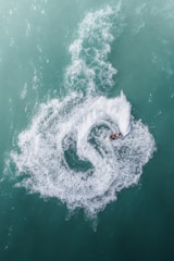 A person riding a jet ski creates a swirling pattern of white water against a backdrop of turquoise sea. The circular trail in the water draws attention, showing a dynamic contrast between the calm water and the swiftly moving vehicle.