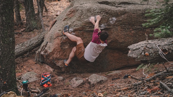 A person wearing a white and maroon shirt and brown shorts is bouldering on a large rock in a forested area. Climbing gear, including a crash pad and water bottle, is scattered nearby on the forest floor, which is covered with pine needles and fallen branches. The environment is lush and green with trees surrounding the area.