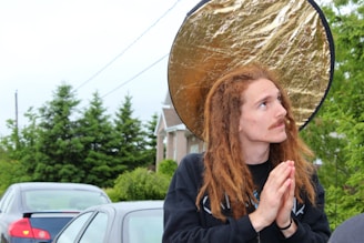A person with long, reddish-brown dreadlocks and a mustache is looking upwards with hands together in a gesture of prayer. Behind the individual is a round, reflective gold disc. The background features parked cars and a suburban setting with green trees and a house visible.