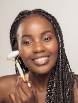 A person with braided hair is smiling while using a facial roller on their cheek. The roller has a white and gold handle, and the background is neutral.