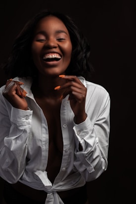 A person is laughing joyfully while facing the camera. They are wearing an unbuttoned white shirt and have short, dark hair. Their fingernails are painted in a bright orange color.