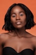 A portrait of a woman with dark hair styled in a bob, wearing a strapless black top against an orange background. Her expression is calm and poised, with soft lighting enhancing her features.