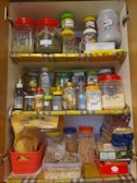 An organized kitchen cabinet filled with various labeled jars containing spices, grains, and other ingredients. The shelves are lined with colorful paper, and the containers are made of plastic and glass, featuring different shapes and sizes. There are also sealed bags of pasta and coffee packets neatly stacked.