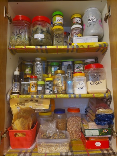 An organized kitchen cabinet filled with various labeled jars containing spices, grains, and other ingredients. The shelves are lined with colorful paper, and the containers are made of plastic and glass, featuring different shapes and sizes. There are also sealed bags of pasta and coffee packets neatly stacked.