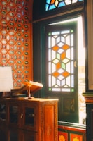An intricately designed interior featuring a stained glass window with geometric patterns, illuminated by sunlight. The wall is adorned with colorful hexagonal tiles in red and blue. Furniture includes a carved wooden cabinet and a wooden stand holding what appears to be books or documents.