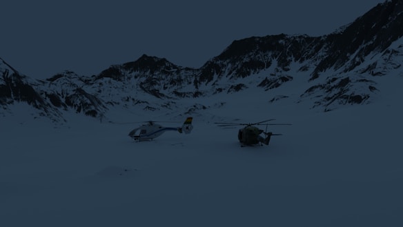Two helicopters are positioned on a snow-covered landscape surrounded by mountain peaks. The sky appears overcast, with the terrain and helicopters being partially obscured by the shadows.