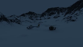 Two helicopters are positioned on a snow-covered landscape surrounded by mountain peaks. The sky appears overcast, with the terrain and helicopters being partially obscured by the shadows.