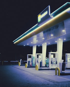 A dimly lit gas station at night with glowing neon lights under the canopy. Several fuel pumps are visible, and two individuals are near one of the pumps. The surrounding area is dark and the cobblestone pavement is visible under the lights.