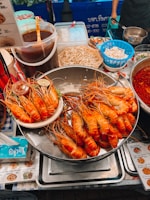 A bustling street food stall displaying a large tray piled with grilled prawns. Nearby are containers of sauces, raw bean sprouts, and other ingredients. The area is vibrant with a mix of various food items and utensils, and there are price lists and signs in a foreign script.