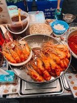 A bustling street food stall displaying a large tray piled with grilled prawns. Nearby are containers of sauces, raw bean sprouts, and other ingredients. The area is vibrant with a mix of various food items and utensils, and there are price lists and signs in a foreign script.