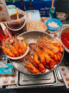 A bustling street food stall displaying a large tray piled with grilled prawns. Nearby are containers of sauces, raw bean sprouts, and other ingredients. The area is vibrant with a mix of various food items and utensils, and there are price lists and signs in a foreign script.