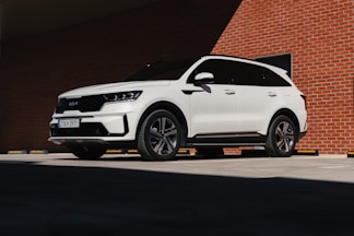 A white SUV is parked in a shadowed area against a backdrop of a red brick wall. Sunlight partially illuminates the upper part of the vehicle, highlighting its sleek design and modern features. The license plate is visible, and the wheels are turned slightly to the left.