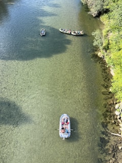 Several inflatable boats with people are floating gently on a clear, shallow river. The water is transparent, revealing the rocky riverbed beneath. Lush greenery lines the riverbank, adding a touch of nature to the serene setting.