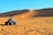 A person rides an ATV across expansive sand dunes with tire tracks visible on the ground. The landscape is characterized by smooth, rolling sand hills under a clear blue sky.