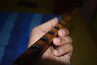 A person is holding a wooden flute with several finger holes visible. The hand is positioned to play, and the background is blurred with shades of blue.