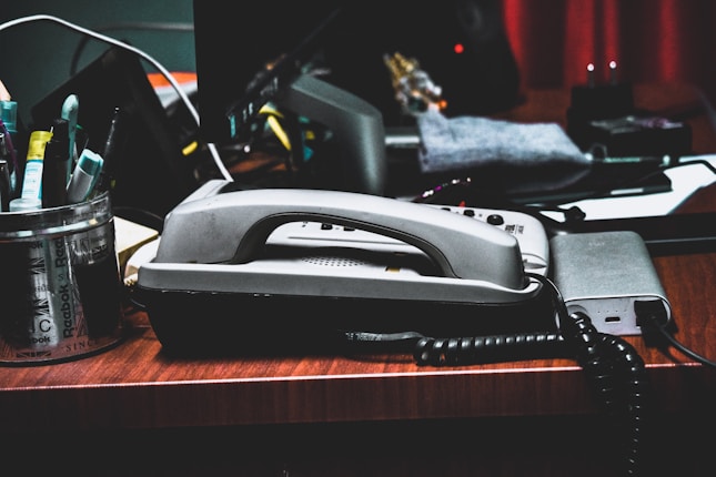 A cluttered desk features an old-fashioned corded telephone at the center. Nearby, a metal container holds various pens and markers. The desk also has cables, a connected electronic device, and some papers scattered around. The ambient lighting suggests a dimly lit room.